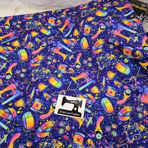 Puede incluir: Una tela azul con un patrón repetitivo colorido de máquinas de coser, carretes de hilo y tijeras. La tela está impresa con un efecto de pintura salpicada. El texto "Rubyjam Fabric" está impreso en una etiqueta blanca.