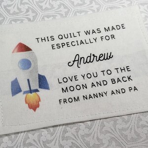 May include: White fabric label with text and a rocket ship illustration. The text reads "THIS QUILT WAS MADE ESPECIALLY FOR Andrew LOVE YOU TO THE MOON AND BACK FROM NANNY AND PA."