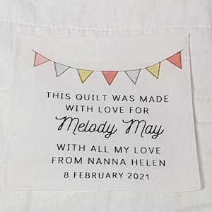 May include: A white fabric label with a colourful bunting design at the top. The text reads "THIS QUILT WAS MADE WITH LOVE FOR Melody May WITH ALL MY LOVE FROM NANNA HELEN 8 FEBRUARY 2021".