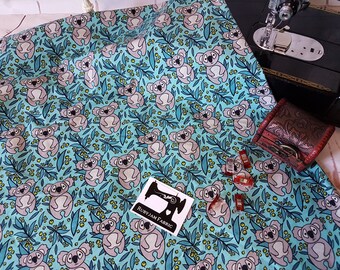 May include: Teal fabric with a repeating pattern of koalas and eucalyptus leaves. The fabric is printed with a light gray koala design and yellow flowers. The text "Rubyjam Fabric" is printed on a white tag.