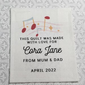 Könnte beinhalten: Ein weißes Stofflabel mit einem Musiknoten-Design und dem Text "THIS QUILT WAS MADE WITH LOVE FOR Cora Jane FROM MUM & DAD APRIL 2022".