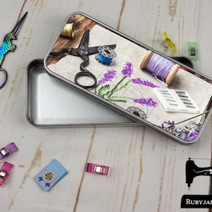 Puede incluir: Una lata de metal plateada con un diseño floral en la tapa. El diseño presenta flores de lavanda, un carrete de hilo, tijeras y agujas. La lata está abierta y el interior está vacío.