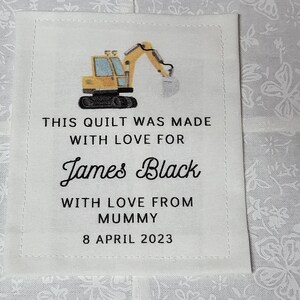 Pode incluir: Um rótulo de tecido branco com texto preto que diz "This quilt was made with love for James Black with love from Mummy 8 April 2023". Um brinquedo escavador amarelo é mostrado acima do texto.