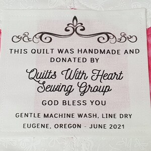 Puede incluir: Etiqueta de tela blanca con texto negro que dice "This quilt was handmade and donated by Quilts With Heart Sewing Group. God bless you. Gentle machine wash, line dry. Eugene, Oregon - June 2021."