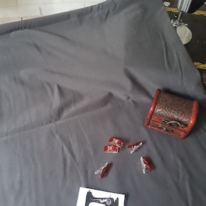 Puede incluir: Un telón de fondo de tela gris con una pequeña caja de madera, clips de costura rojos y una pegatina blanca con un gráfico de máquina de coser y el texto "Rubyjam Fabric".