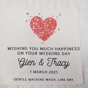 May include: A white fabric wedding favour tag with a red heart made of butterflies. The tag says "Wishing you much happiness on your wedding day Glen & Tracy 1 March 2021 Gentle machine wash, line dry."
