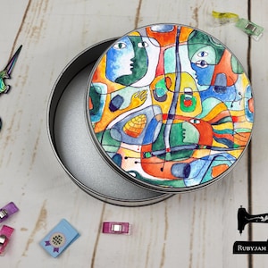 Puede incluir: Una lata redonda plateada con una tapa que presenta un diseño abstracto de acuarela en tonos azules, naranjas y verdes. La imagen también incluye unas tijeras de unicornio arcoíris, pinzas de costura y una pequeña tarjeta azul.