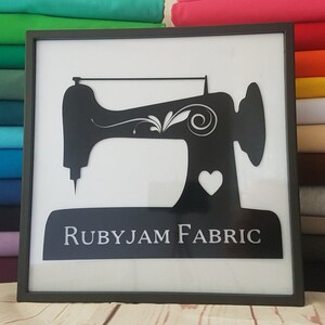 May include: Black and white silhouette of a sewing machine with a heart and the text "RUBYJAM FABRIC" on a white background in a black frame.