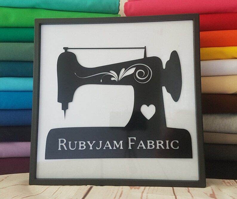 May include: Black and white silhouette of a sewing machine with a heart and the text "RUBYJAM FABRIC" on a white background in a black frame.