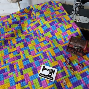 以下が含まれることがあります： カラフルなレゴブロックの繰り返しパターンが施された生地。生地は白い表面に置かれ、背景にはミシンと小さな木製の箱があります。生地には「RubyJam Fabric」と書かれたラベルが付いています。
