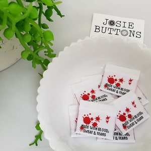 Puede incluir: Un tazón blanco lleno de pequeñas etiquetas de tela blanca con salpicaduras de pintura roja y el texto "Made with Blood, Sweat & Tears".