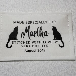 May include: White fabric label with black stitching and text. The text reads "Made especially for Martha, Stitched with love by Vera Biefield, August 2019". Two black cat silhouettes flank the name Martha.