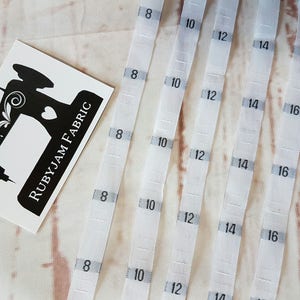 Puede incluir: Cinco cintas métricas de tela blanca con números negros impresos en ellas. Los números en las cintas son 8, 10, 12, 14 y 16. Las cintas están dispuestas en una fila sobre una superficie de madera marrón claro.