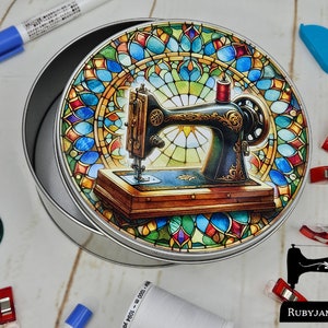 Puede incluir: Una lata redonda de metal con un diseño de vitral colorido que presenta una máquina de coser vintage. La lata es plateada con un fondo blanco y la máquina de coser es negra con un carrete de hilo rojo. El diseño está rodeado por un borde de vitral colorido.