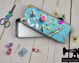 May include: A metal tin with a blue background and a variety of sewing supplies, including buttons, thread, needles, and a measuring tape. The tin is open and the inside is silver.