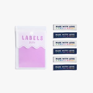 Puede incluir: Un conjunto de cinco etiquetas termoadhesivas en blanco y azul marino con el texto "Made with Love + Swear Words" en colores arcoíris. Las etiquetas están empaquetadas en una bolsa de plástico transparente con una etiqueta rosa que dice "Labels by Katm".