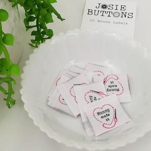 Puede incluir: Un cuenco blanco con borde festoneado contiene varias etiquetas tejidas cuadradas pequeñas. Cada etiqueta presenta un diseño de corazón con una aguja e hilo, y el texto "Mummy made it" en rosa. Una pequeña tarjeta dice "JOSIE BUTTONS 10 Woven Labels".
