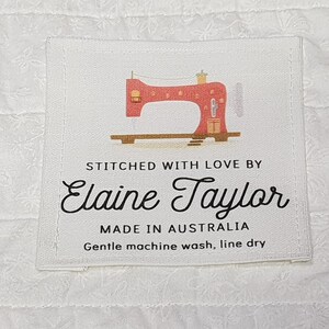 May include: A white fabric label featuring a red sewing machine illustration. Text reads "STITCHED WITH LOVE BY Elaine Taylor MADE IN AUSTRALIA Gentle machine wash, line dry." The label is set against a white patterned backdrop.