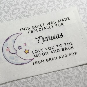 May include: A white fabric label with a blue and purple crescent moon and stars. The text on the label reads "This quilt was made especially for Nicholas Love you to the moon and back From Gran and Pop."