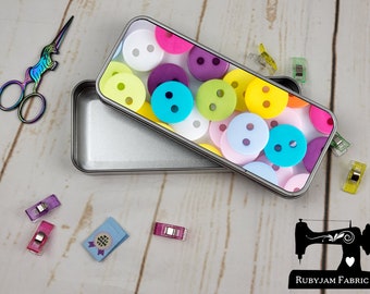 May include: A silver metal tin with a colorful button design on the lid. The tin is open and the inside is empty. The tin is on a white wooden surface.