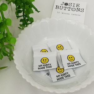Puede incluir: Etiquetas tejidas blancas con una carita sonriente amarilla y el texto "MY AUNTY MADE THIS". Las etiquetas están en un plato blanco con volantes, con una etiqueta "JOSIE BUTTONS". Planta verde en el fondo.