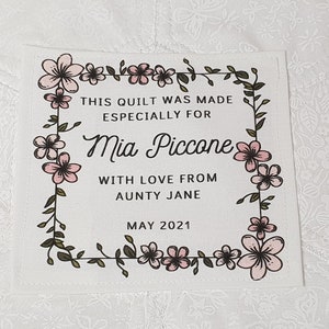 May include: A white fabric quilt label with a floral border. The label reads "This quilt was made especially for Mia Piccone with love from Aunty Jane May 2021".