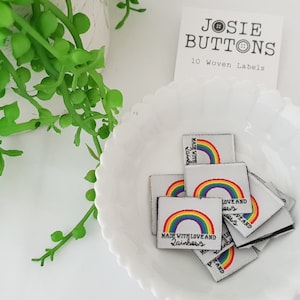 Puede incluir: Un conjunto de 10 etiquetas tejidas blancas con un arcoíris y el texto "Made with Love and Rainbows".