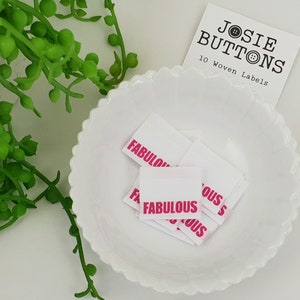 Puede incluir: Un bol blanco lleno de diez etiquetas tejidas blancas con la palabra "FABULOUS" impresa en rosa. Las etiquetas son para ropa o manualidades. Una tarjeta de visita con el texto "JOSIE BUTTONS 10 Woven Labels" está en el fondo.