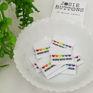 Puede incluir: Un conjunto de 10 etiquetas tejidas blancas con corazones arcoíris y el texto "Worn with Pride".