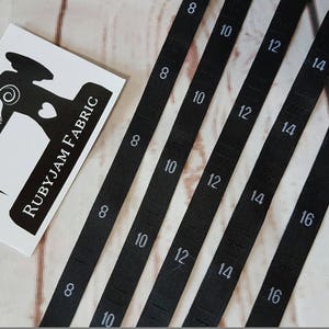 Puede incluir: Cinco cintas métricas de tela negra con números blancos impresos en ellas. Los números en las cintas son 8, 10, 12, 14 y 16.