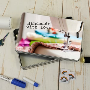 Puede incluir: Una caja de costura de metal plateado con un diseño de tela colorida y el texto "Handmade with love". La caja está abierta y el interior es visible. Hay artículos de costura en la mesa junto a la caja, incluyendo un carrete de hilo, un par de tijeras y un marcador.