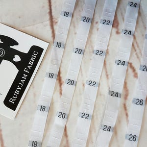 Puede incluir: Cinta métrica de tela blanca con números negros del 18 al 26 pulgadas. La cinta está doblada por la mitad y los números son visibles en ambos lados.