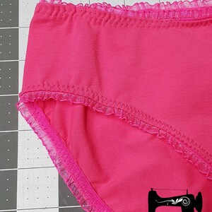 May include: Pink underwear with a lace trim. The underwear is on a gray cutting mat with a sewing machine logo and the text "Rubyjam Fabric".
