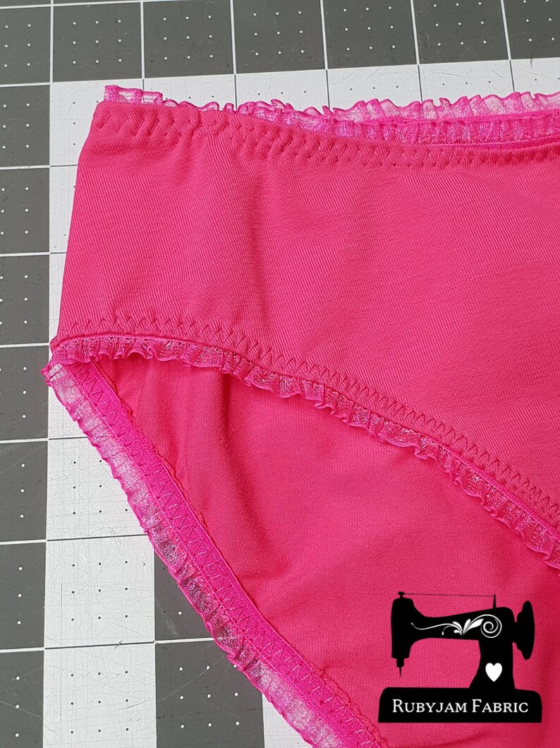 May include: Pink underwear with a lace trim. The underwear is on a gray cutting mat with a sewing machine logo and the text "Rubyjam Fabric".