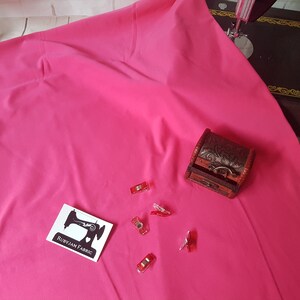 May include: A bright pink fabric with a small wooden box and red sewing clips. The fabric is smooth and has a slight sheen. The box is closed and has a decorative design. The clips are scattered on the fabric.