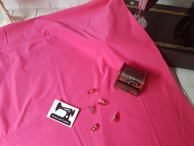 May include: A bright pink fabric with a small wooden box and red sewing clips. The fabric is smooth and has a slight sheen. The box is closed and has a decorative design. The clips are scattered on the fabric.