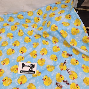 Puede incluir: Tela azul con un patrón repetido de patos de goma amarillos. Los patos están en varias posiciones y algunos tienen un pico rojo. La tela tiene un fondo blanco con un patrón de remolino azul claro.