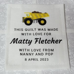 Puede incluir: Una etiqueta de tela blanca con texto negro que dice "This quilt was made with love for Matty Fletcher with love from Nanny and Pop 8 April 2023". Un camión de volteo de juguete amarillo con ruedas negras está representado encima del texto.