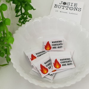 Puede incluir: Pequeñas etiquetas tejidas blancas con el texto "WARNING: CONTENTS ARE HOT!" y un gráfico de llama. Las etiquetas están en un cuenco blanco con volantes, con una etiqueta "Josie Buttons" en el fondo.