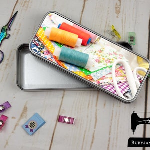 Puede incluir: Una lata de metal plateada con un diseño de tela colorida en la tapa. La tapa presenta carretes de hilo coloridos, recortes de tela y herramientas de costura. La lata está abierta y el interior está vacío. El texto "Rubyjam Fabric" es visible en la esquina inferior derecha de la imagen.