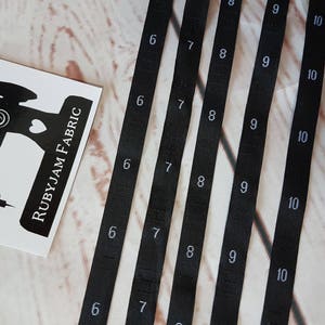 Puede incluir: Seis carretes de cinta de tela negra con números blancos impresos en ellos. Los números en los carretes son 6, 7, 8, 9 y 10. Los carretes están dispuestos en una fila, con los números aumentando de izquierda a derecha.