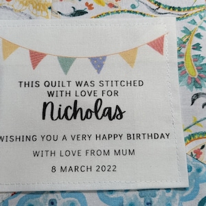 May include: A white fabric label with a colourful bunting design at the top. The text reads "This quilt was stitched with love for Nicholas Wishing you a very happy birthday with love from Mum 8 March 2022".