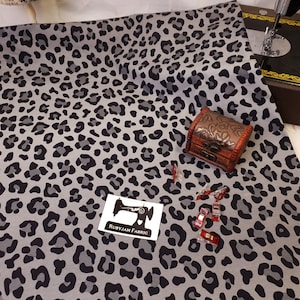 May include: Grey fabric with a black leopard print pattern. The fabric is displayed on a white surface with a small wooden box and sewing clips.