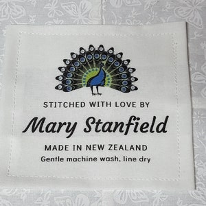 Puede incluir: Etiqueta de tela blanca con un diseño de pavo real y el texto "STITCHED WITH LOVE BY Mary Stanfield MADE IN NEW ZEALAND Gentle machine wash, line dry"