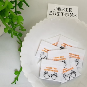 Puede incluir: Un fondo blanco con una pila de etiquetas de tela blanca con el texto "Hice esto con mis manos" y un icono de mano en naranja y negro.