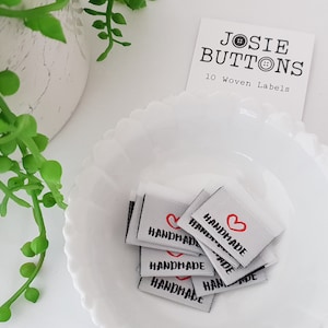 Puede incluir: Un montón de etiquetas cuadradas tejidas con la palabra "HANDMADE" en texto negro y un corazón rojo. Las etiquetas están en un cuenco blanco con volantes. Una tarjeta dice "JOSIE BUTTONS" y "10 Woven Labels". Hay tallos de plantas verdes en el fondo.
