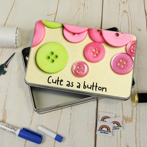 Puede incluir: Una caja metálica con tapa decorada con botones rosas y verdes, con la frase "Cute as a button". La caja está abierta, mostrando su interior. Suministros de costura, como tijeras, hilo y etiquetas, están colocados alrededor.