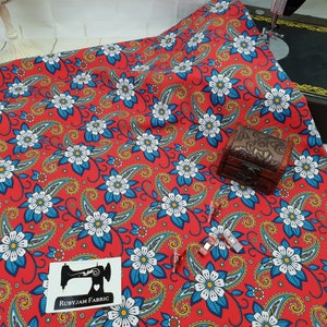 Puede incluir: Tela roja con estampado paisley y floral. La tela presenta flores blancas con centros azules y detalles amarillos. La tela está rodeada por una máquina de coser y una pequeña caja de madera.