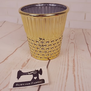 Puede incluir: Un recipiente cilíndrico dorado con una superficie texturizada. El recipiente tiene un diseño acanalado en la parte superior y un patrón punteado en la parte inferior. Una pegatina con un gráfico de máquina de coser y el texto "RUBYJAM FABRIC" es visible.
