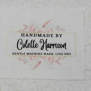 May include: White fabric label with a floral design in pink and grey. The label says "Handmade by Colette Harrison" in black text with a gold rectangle behind the name. The label also says "Gentle Machine Wash, Line Dry" in black text.